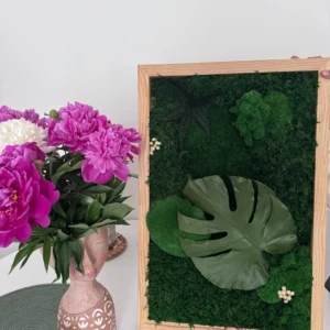 Moosbild mit Monstera-Blatt im Holzrahmen, aufgestellt neben einer Vase mit pinken Pfingstrosen.